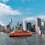 An orange ferry sails past a city on the water.